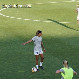 Athletes are on a soccer field engaging in practice. A player wearing a jersey with the number 14 is standing with a soccer ball at their feet, pointing to the right. Teammates are scattered around the field, some stretching or preparing for drills. A coach or staff member is carrying equipment, possibly looking towards the players.