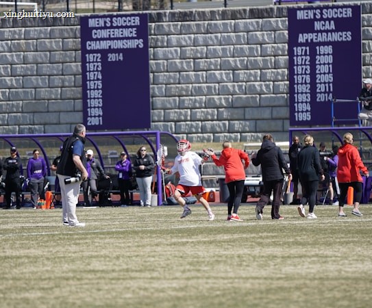 A sports team is practicing or playing on a grassy field in front of a large stone wall with banners displaying men's soccer achievements. People are wearing sports attire, and some are engaged in conversation or coaching.